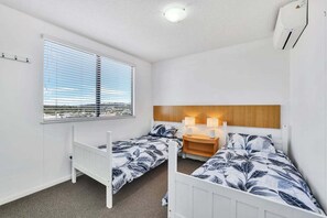 A room with two single beds, a window with blinds, a bedside table with lamps, and an air conditioning unit. - Amazing Ocean Views from Unit 87-Catalina-II (Maroochydore)