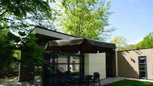 A modern outdoor patio with a glass enclosure, a table with chairs, and a large umbrella. - Holiday Home in Kaatsheuvel With Terrace (Kaatsheuvel)