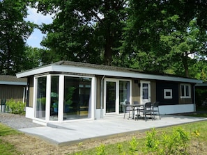 Ein modernes Haus mit einer gläsernen Veranda, Sitzgelegenheiten im Freien und einem großen Baum im Hintergrund.