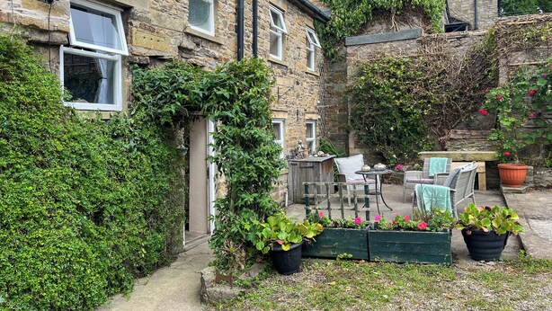 Un bâtiment en pierre dont les murs sont recouverts de lierre, un petit patio avec des meubles d’extérieur et des plantes en pot.