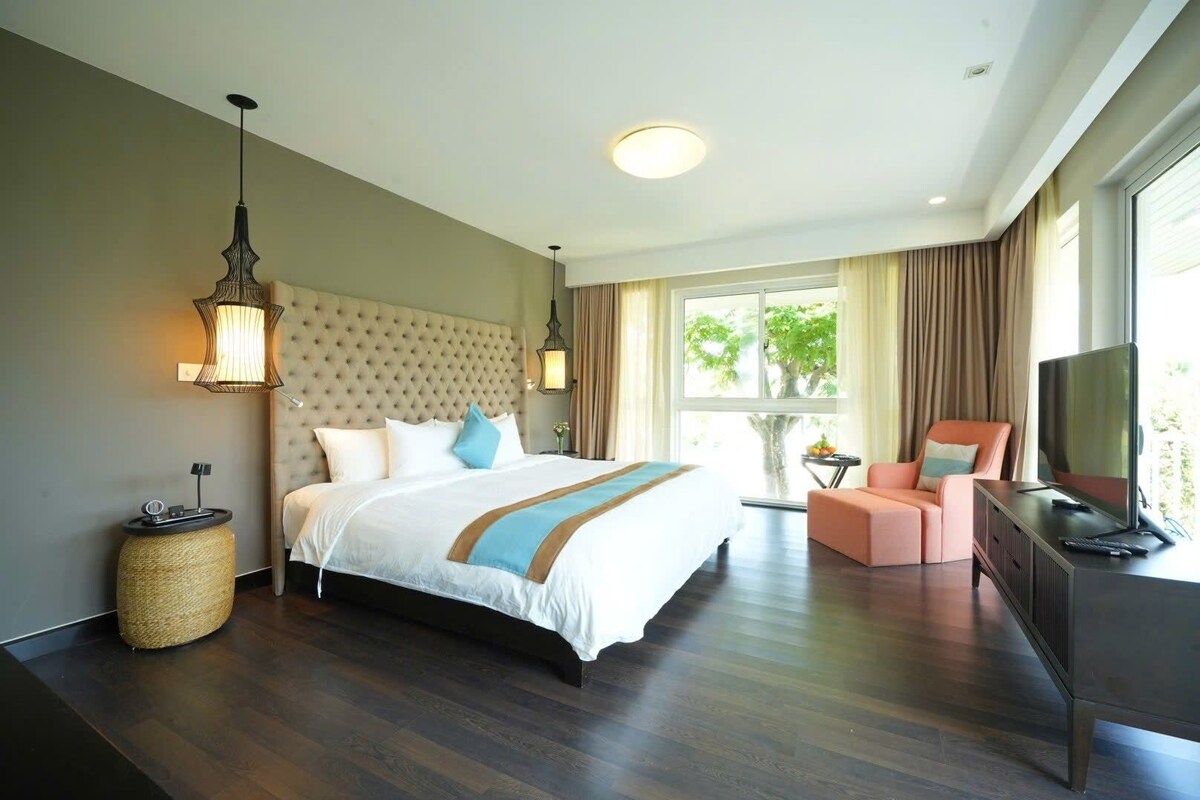 A hotel room with a large bed, a television, a seating area, and a window with a view of trees.