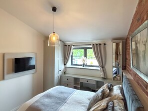 Quarto com cama, televisão fixada na parede, luminária pendente, janela com cortinas e parede de tijolos.