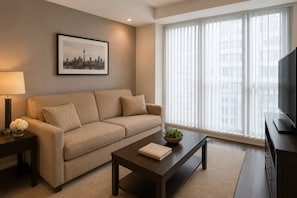 A modern living room with a sofa, a coffee table, a TV, and a window with blinds. - Solenne Park Hotel (Toronto)