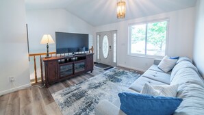 Un salon doté d’un téléviseur à écran plat, d’un meuble de rangement en bois, d’un tapis à motifs, d’un canapé avec des coussins et d’une grande fenêtre.