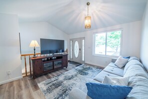 Ein Wohnzimmer mit Flachbildfernseher, einer Holzunterhaltungsecke, einem gemusterten Teppich, einer Couch mit Kissen und einem großen Fenster.