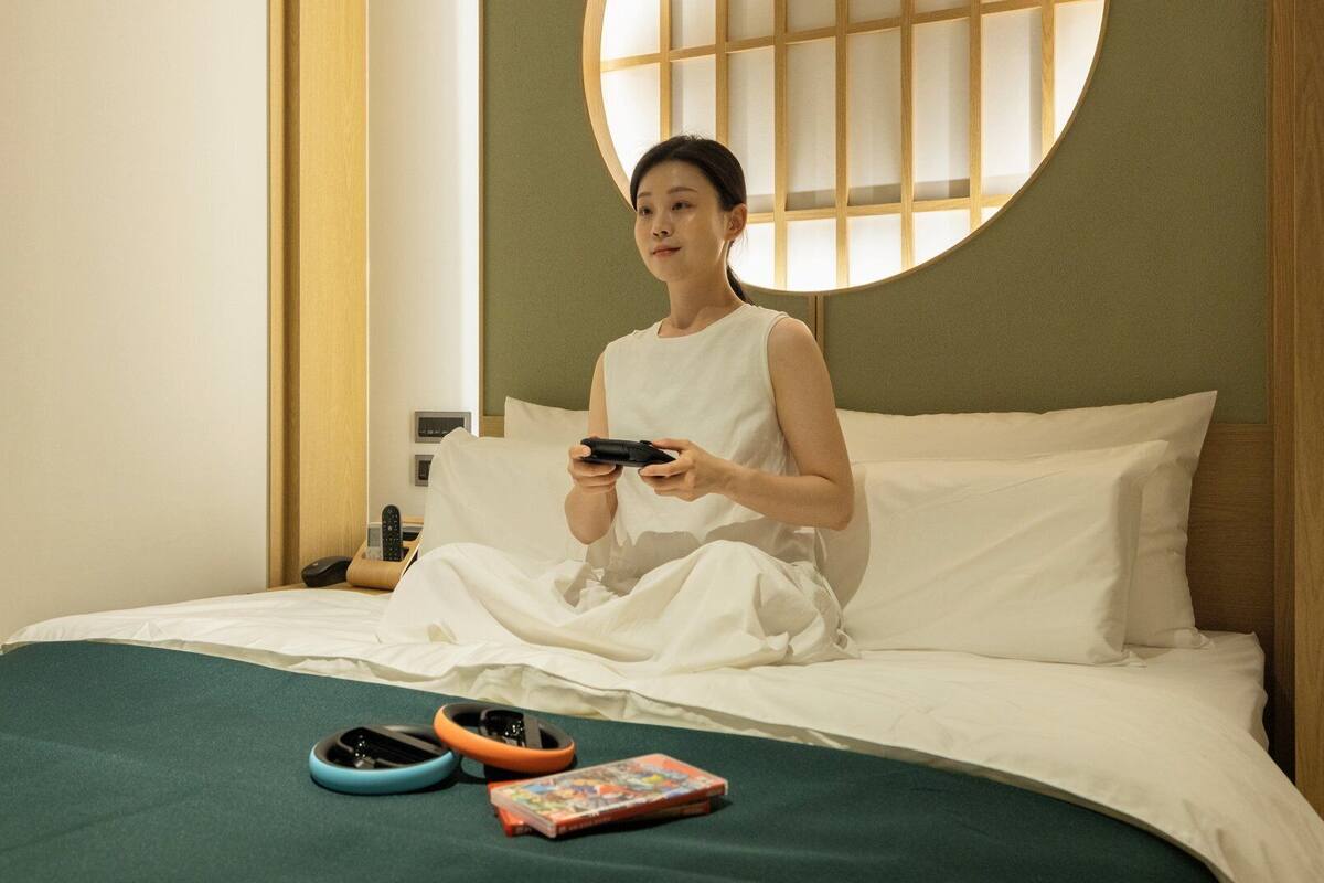 A person sitting on a bed in a room with a round window, holding a game controller.