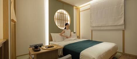 A hotel room with a bed, a desk with a remote control, a chair, and a round window.