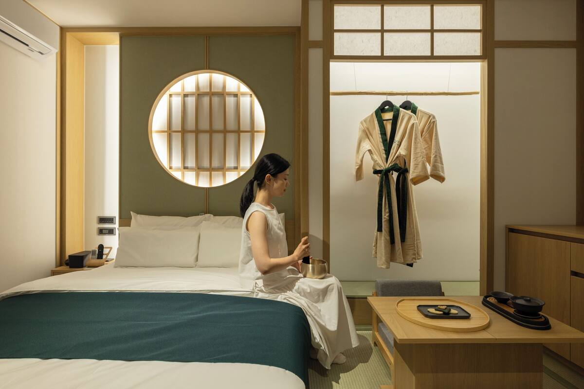 A hotel room with a bed, a round window, a table with a bowl and a tray, and a coat hanging on the wall.