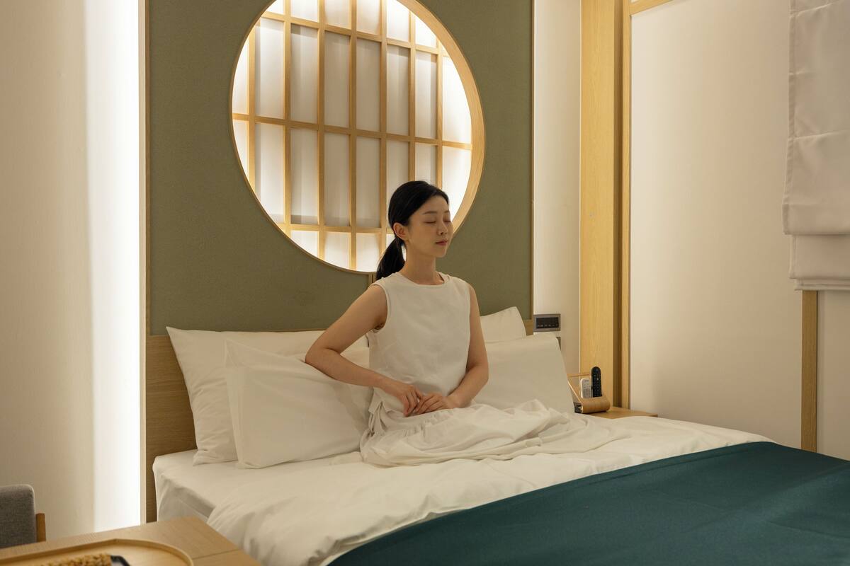 A hotel room with a bed, pillows, a bedside table with a phone, and a wall with a circular window.