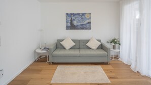 Un salon moderne avec un canapé gris, deux coussins décoratifs blancs, un tableau représentant un ciel nocturne, une petite table avec un livre et une bougie, et un tapis blanc sur le sol.