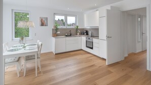 Une cuisine moderne avec des meubles blancs, une table à manger avec des chaises et une vue sur l’extérieur grâce à de grandes fenêtres.