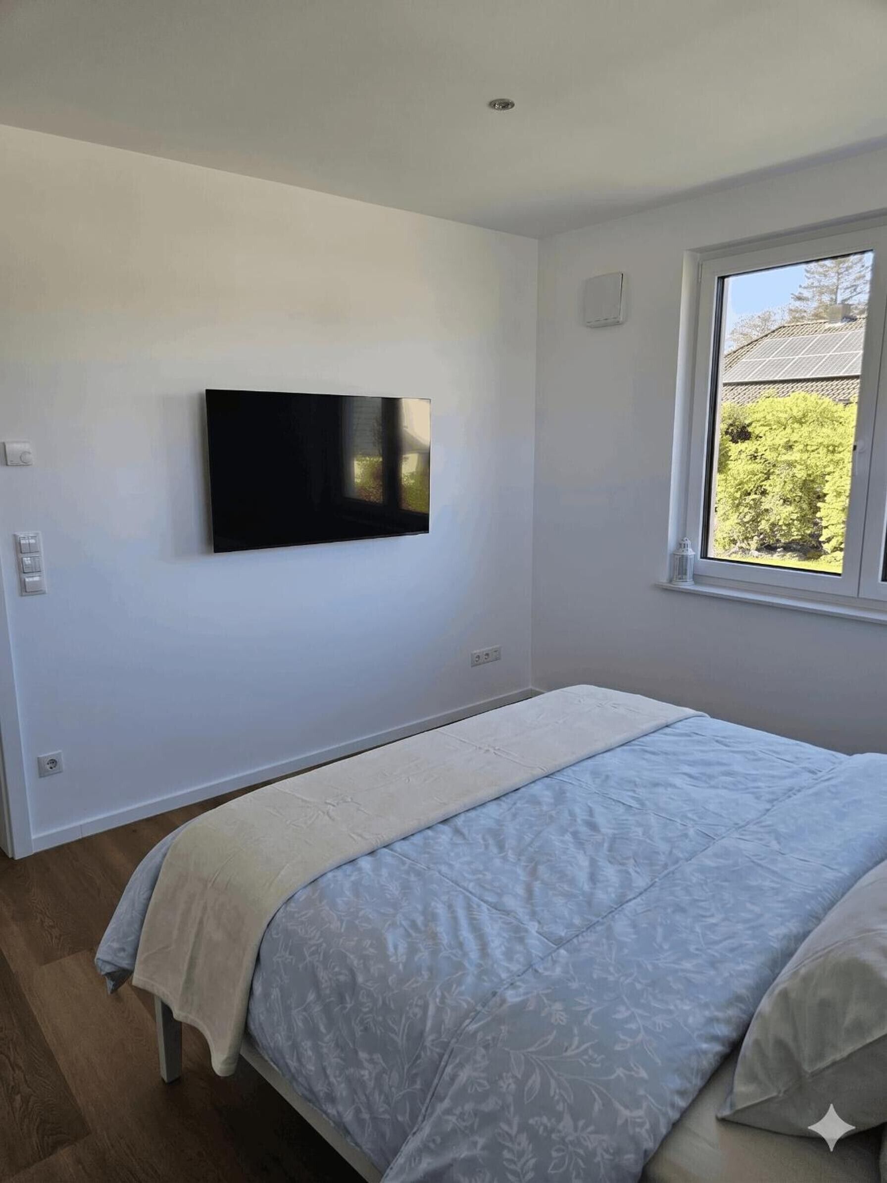 Ein Schlafzimmer mit Bett, einem an der Wand befestigten Fernseher, einem Fenster mit Aussicht und einem Thermostat.