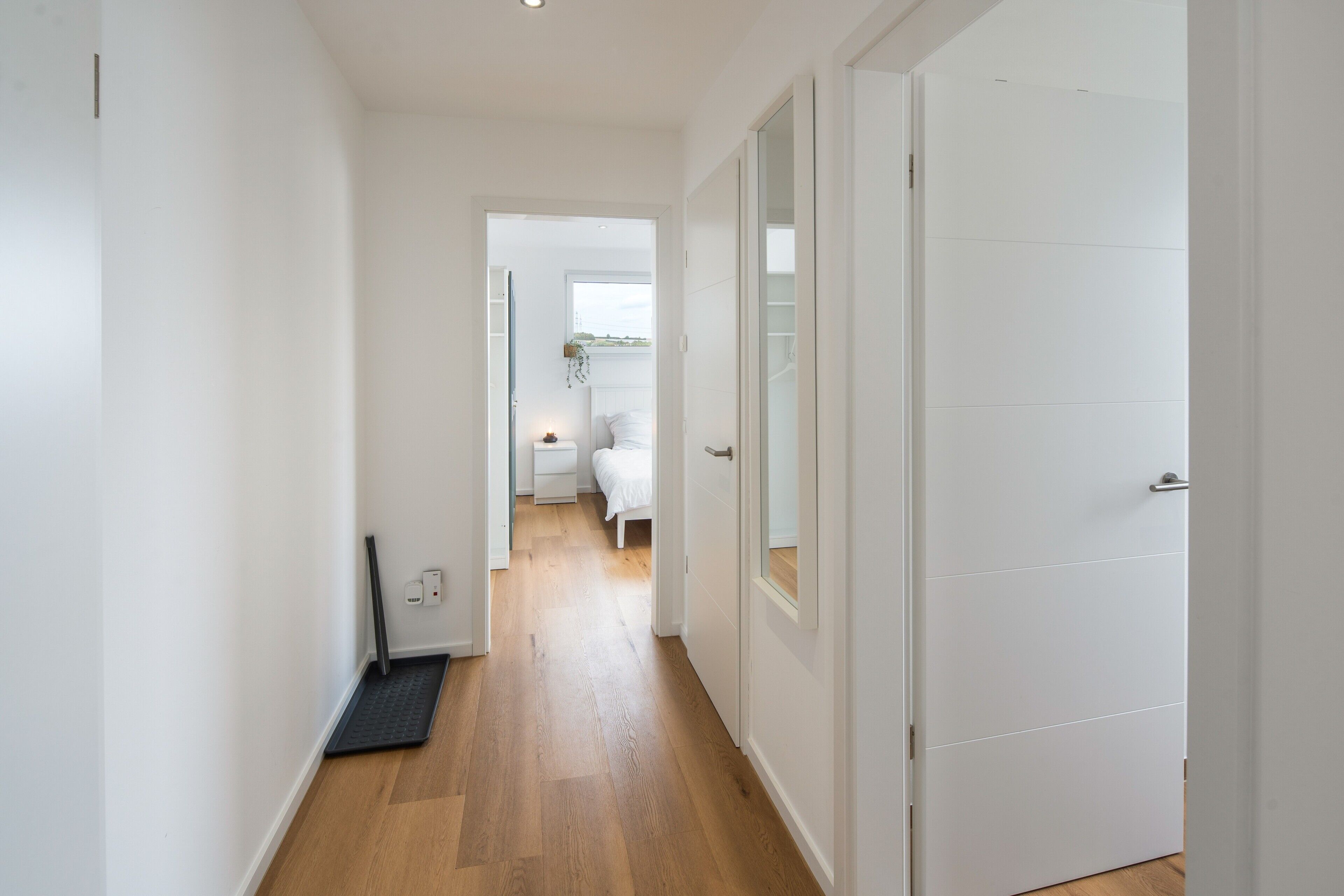 Un pasillo con piso de madera, paredes blancas y una puerta que da a un dormitorio con cama y ventana.