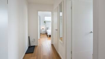 Un pasillo con piso de madera, paredes blancas y una puerta que da a un dormitorio con cama y ventana.