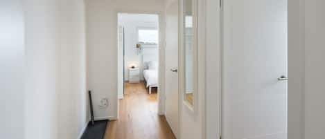 Un pasillo con piso de madera, paredes blancas y una puerta que da a un dormitorio con cama y ventana.