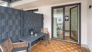 Un área de estar moderna al aire libre con una mesa, sillas, piso de madera y una puerta de vidrio que da a un espacio interior.