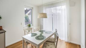 Comedor con una mesa blanca, dos sillas, una lámpara colgante y una ventana que da a una casa.