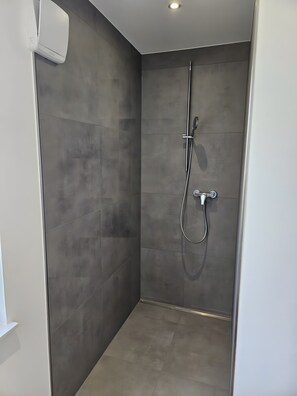 Ducha moderna con cabezal de ducha de mano y grifo empotrado en la pared, ubicada en un rincón con azulejos grises y una luz de techo empotrada.