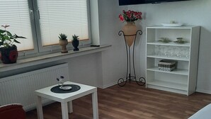 A room with a TV, a small table with a vase, a shelf with decorative items, and a window with blinds. - Vacation Apartment on the Weser River (Elsfleth)
