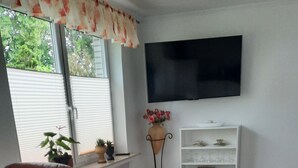 A living room with a red chair, a white table, a TV mounted on the wall, and a window with blinds. - Vacation Apartment on the Weser River (Elsfleth)