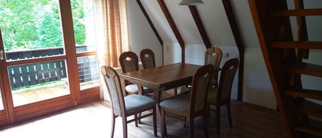 A dining area with a wooden table and chairs, large windows with curtains, and a view of greenery outside.