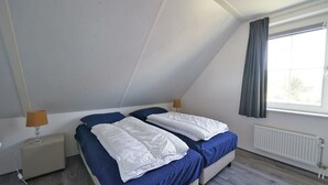Ein Schlafzimmer mit schrägen Wänden, einem Bett mit weißer Bettwäsche, einem Nachttisch mit Lampe und einem Fenster mit Vorhängen.