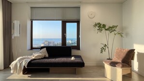 Quarto moderno com cama, mesa de apoio, cadeira, planta e janela com vista para a cidade.