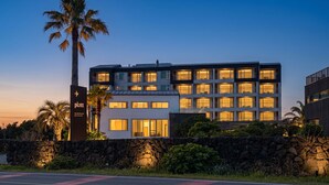 Um hotel de vários andares com entrada em parede de pedra, uma palmeira e uma placa com a inscrição 'plott'.