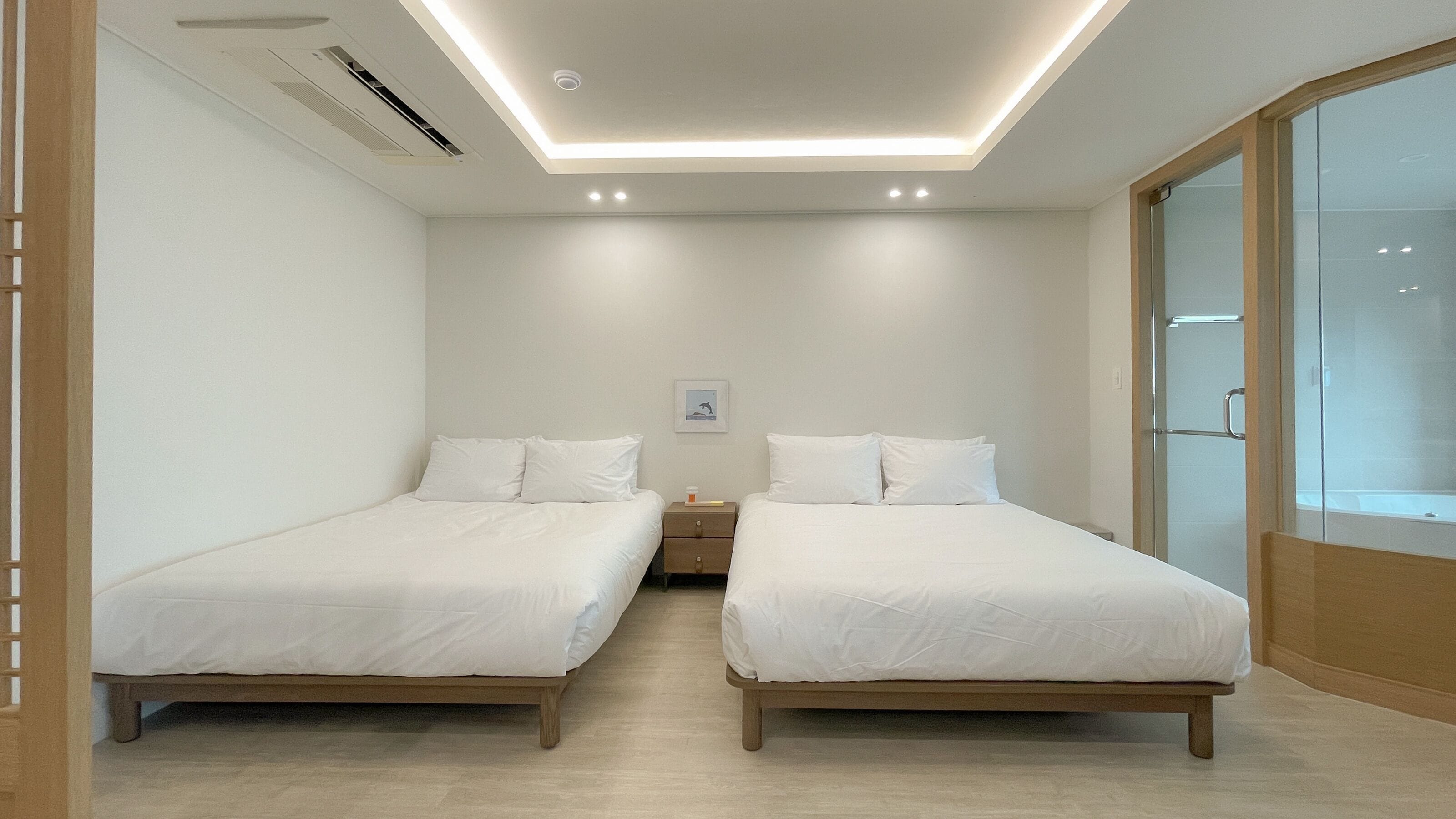A hotel room with two beds, a bedside table, a picture on the wall, and a sliding glass door to the bathroom.