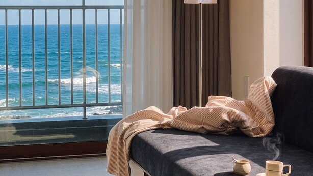 Quarto com cama, mesa de cabeceira com uma chávena e uma caneca, candeeiro de chão e vista para o mar.