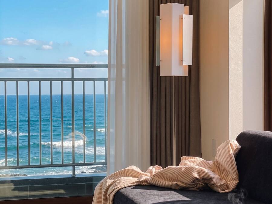 A room with a bed, a bedside table with a cup and a mug, a floor lamp, and a view of the ocean.