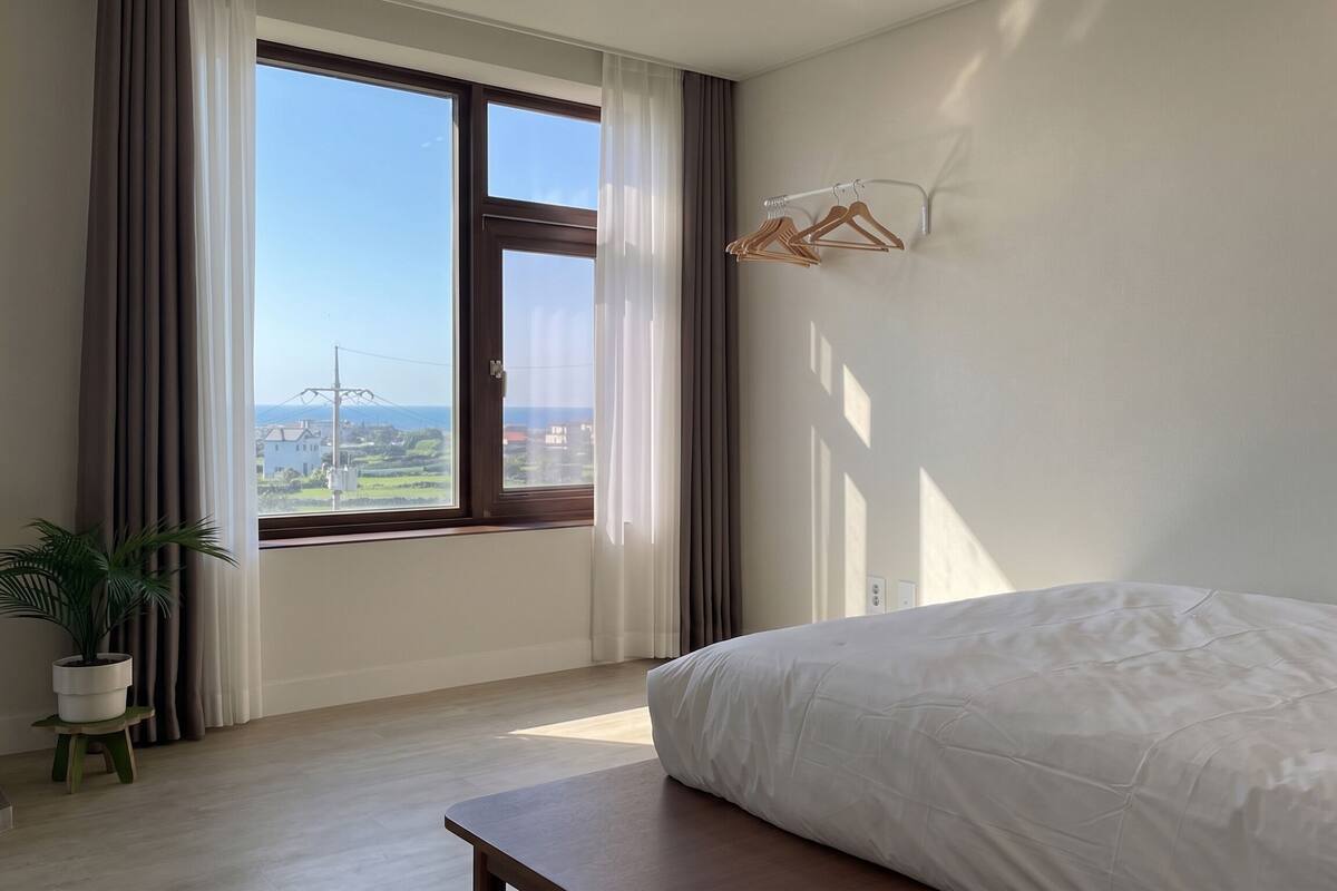 Un dormitorio con un ventanal grande, una cama, una mesita de noche y una lámpara colgante.