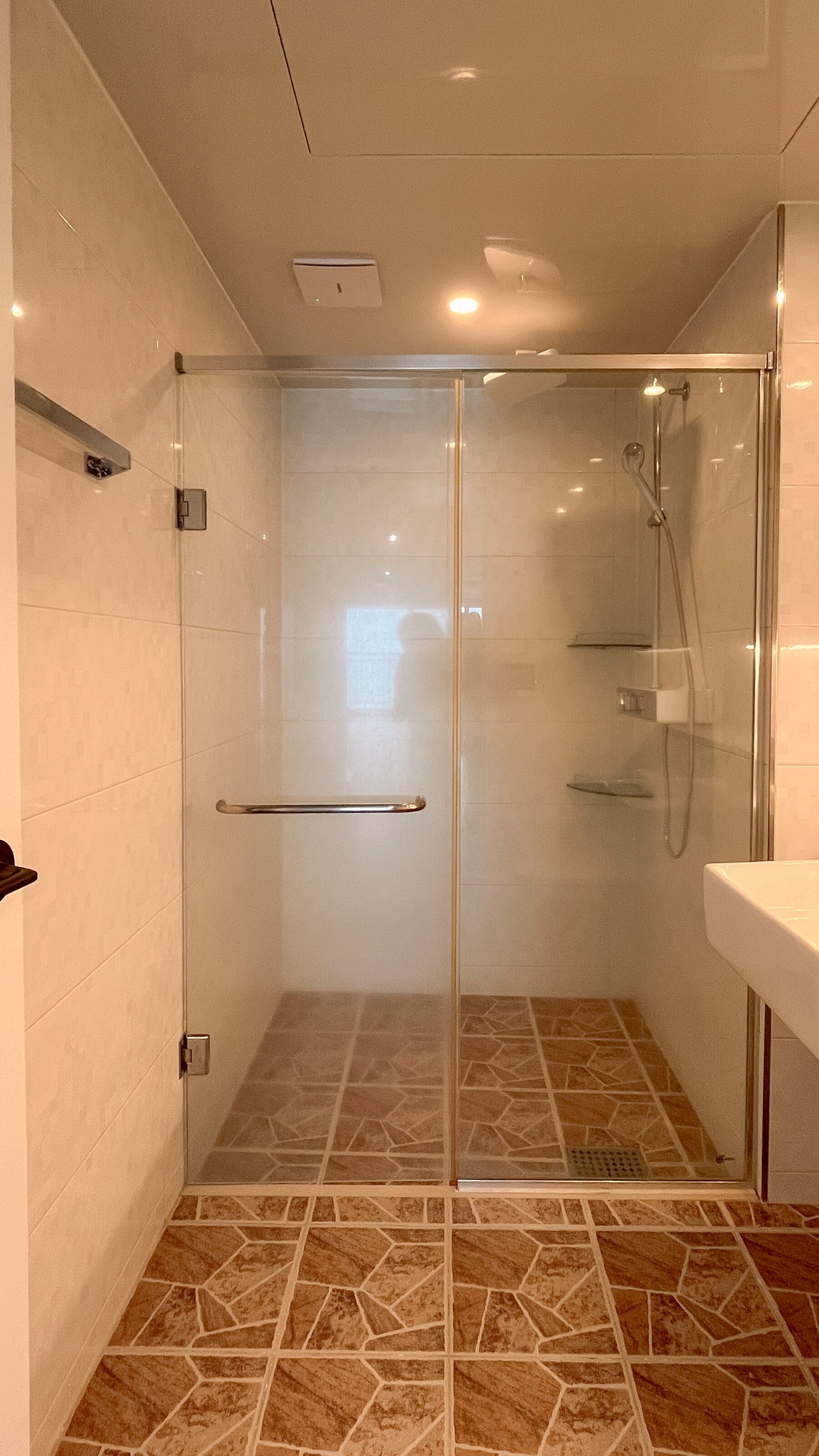 A glass-enclosed shower with a handrail, tiled floor, and a white sink.