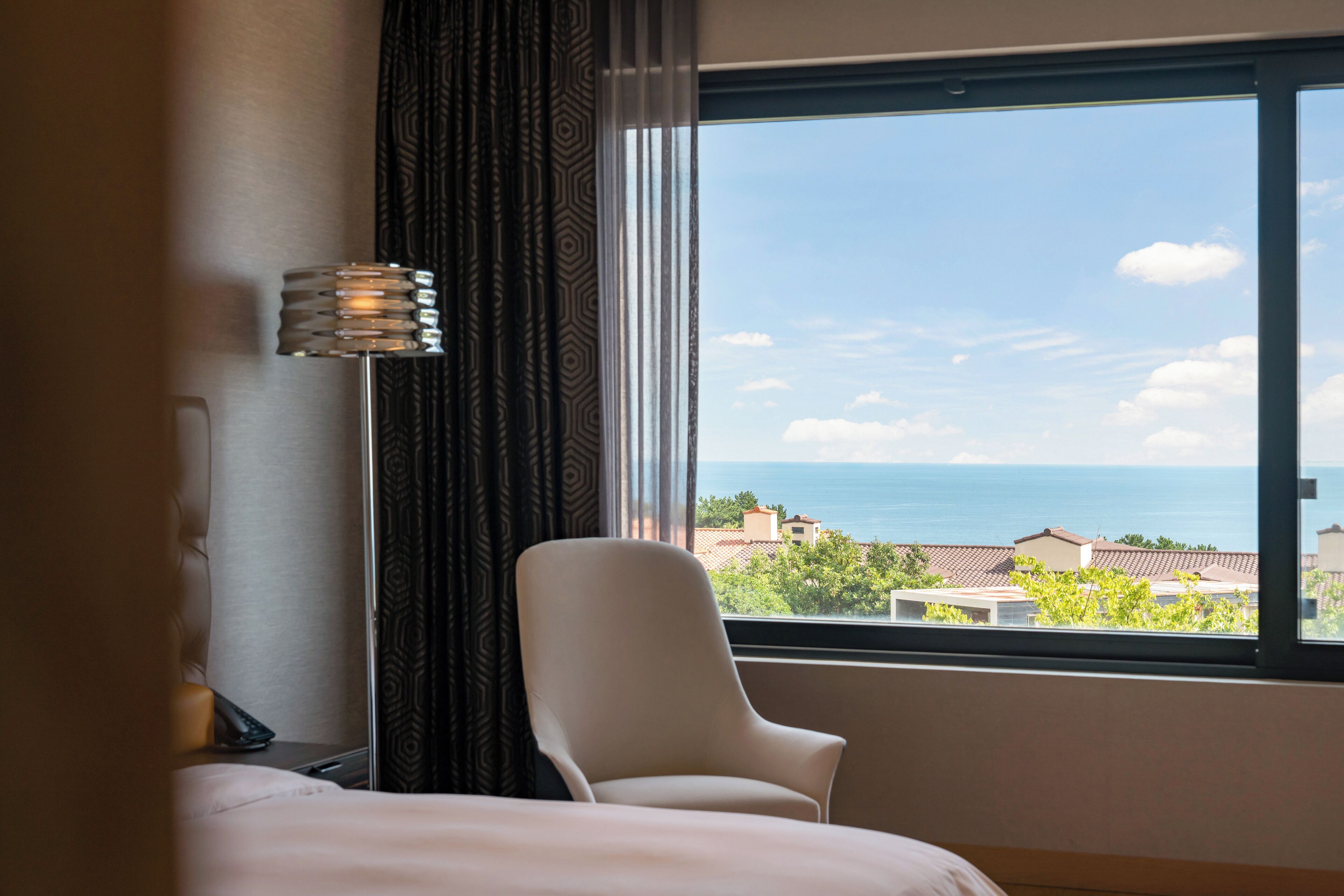 Et hotelværelse med en seng, en stol, en lampe og et vindue med udsigt over havet og bygninger.