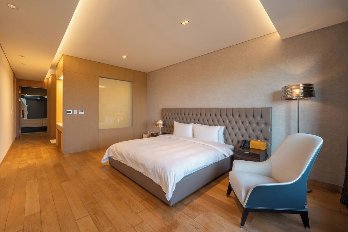 A modern hotel room with a large bed, a chair, a bedside table, a lamp, and a wardrobe.