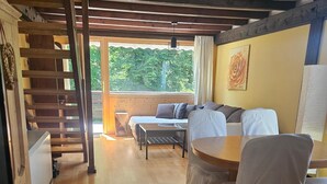 Una habitación con piso de madera, una cama con cabecera, un sofá, una mesa y sillas. Se observa una escalera a la izquierda y una ventana con vista a árboles.