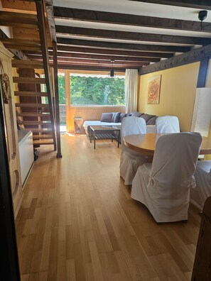 Una habitación con piso de madera, una cama con cabecera, un sofá, una mesa y sillas. Se observa una escalera a la izquierda y una ventana con vista a árboles.