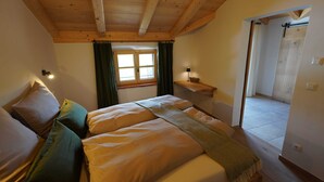 Un dormitori amb sostre de fusta, un llit amb almoades, una tauleta de nit, una finestra amb cortines i una porta que dona a una altra habitació.