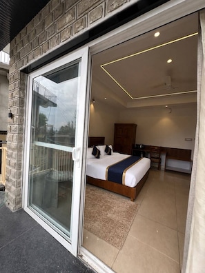 A hotel room with a sliding glass door leading to a balcony, a bed with a headboard, a wooden cabinet, and a small desk with a chair. - Hotel Indradhar (Dharamshala)