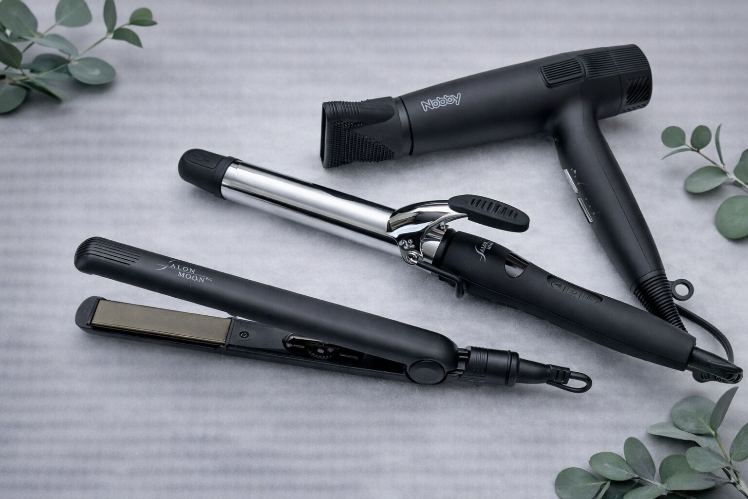 Four hair styling tools on a textured surface with green foliage.