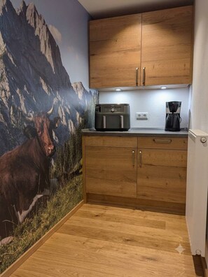A kitchen with wooden cabinets, a coffee maker, and a mountain mural on the wall. - "Auf da Oim" Vacation Rental, 1–2 people, balcony, Wi-Fi, shower/toilet (Ramsau bei Berchtesgaden)