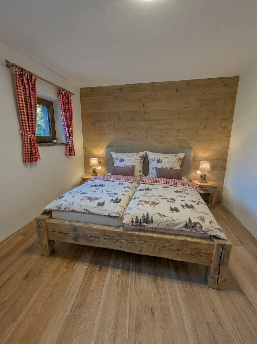 Een houten bed met een geruit sprei, twee nachtkastjes met lampen, en een raam met rode gordijnen.