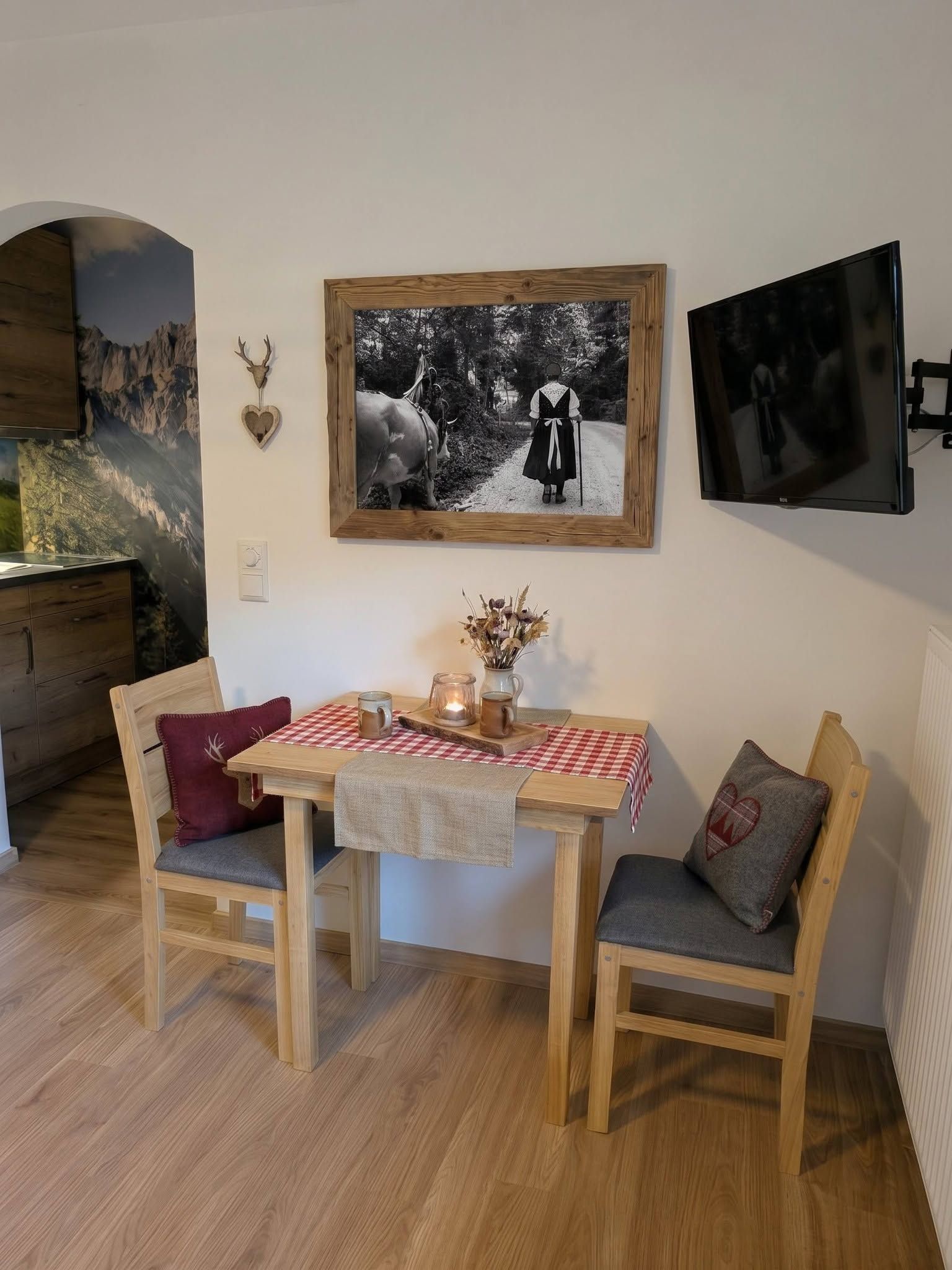 Een eethoek met een houten tafel, twee stoelen, een rood-wit geruit tafelkleed, en een zwart-wit foto in een lijst erboven.