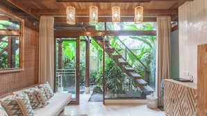 A room with a wooden ceiling, a staircase leading to a balcony, a sofa with patterned cushions, and a cabinet with a geometric design. - Kima Surf Camp Bali Seminyak (Seminyak)