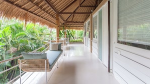 A covered balcony with a thatched roof, a white sofa, and lush green plants. - Kima Surf Camp Bali Seminyak (Seminyak)