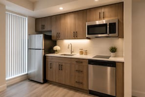 A modern kitchen with wooden cabinets, stainless steel appliances, a dishwasher, and a window with blinds. - The Montclair Suites (Toronto)