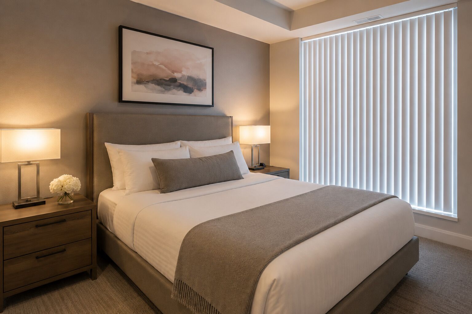 A neatly arranged bedroom with a large bed, two bedside tables, a nightstand, a framed picture on the wall, and a window with blinds.