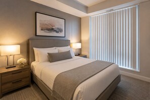 A neatly arranged hotel room with a large bed, two bedside tables, a nightstand, a framed picture on the wall, and a window with blinds. - The Montclair Suites (Toronto)
