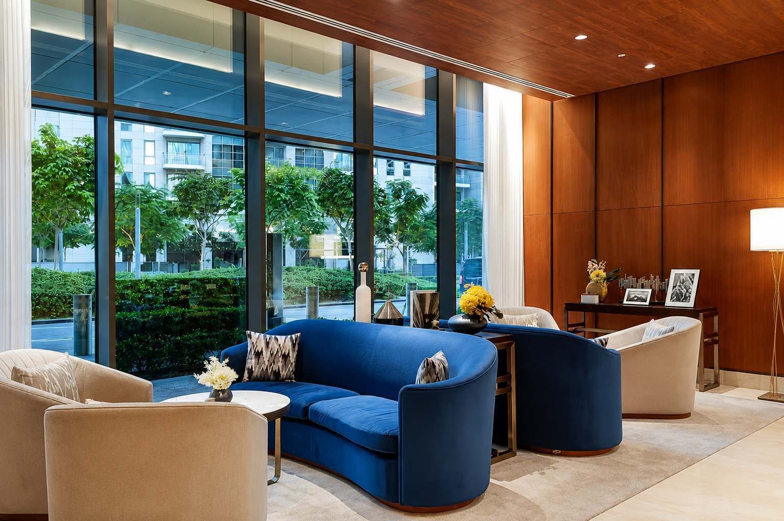 Un moderno vestíbulo de hotel con un sofá azul, sillones beige, una pequeña mesa con un jarrón y un ventanal con vistas a la ciudad.
