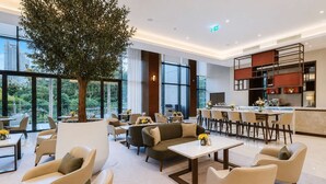 A modern hotel lobby with a large tree, seating areas, a bar, and a kitchen visible in the background. - Address Opera 1BR, Boulevard View-Hotel Style Stay (Dubai)